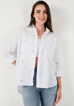 White button-up shirt with light blue heart embroidery, relaxed fit, long sleeves rolled up, and a slightly longer back hem.