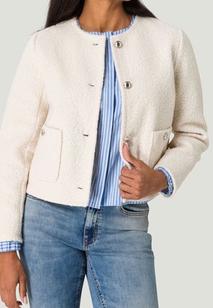Femme portant une veste polaire crème boutonnée sur une chemise rayée bleu et blanc avec un jean bleu.
