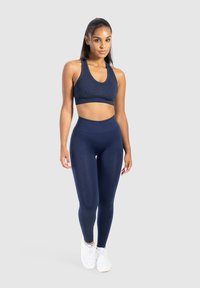 Vrouw in een marineblauwe sportbeha en legging met witte sneakers tegen een effen lichtgrijze achtergrond.