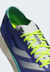 Zapatilla deportiva morada con parte superior texturizada, acentos en blanco y gris, cordones amarillos neón y un interior verde claro. Cuenta con una entresuela ligera.