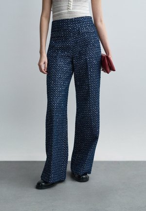 Femme portant un pantalon large taille haute en tissu texturé bleu marine, un haut sans manches blanc avec des boutons, des chaussures noires, tenant une pochette rouge pliée dans la main droite.
