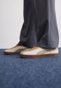 Sneakers in suede beige e oliva con suole in gomma marrone, dotate di sovrapposizione testurizzata, lacci e un accento con logo sul lato.