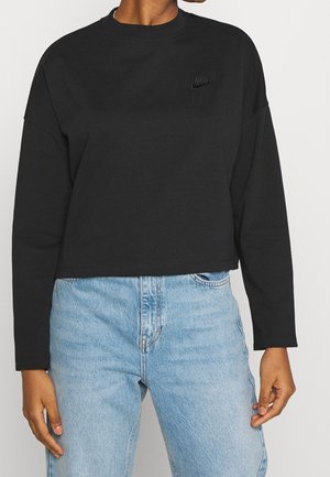 Persoon draagt een zwart cropped Nike-sweatshirt met lange mouwen en lichtblauwe high-waisted jeans tegen een effen achtergrond.