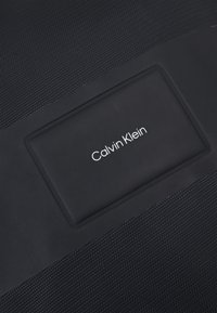 Borsa in tessuto nera Calvin Klein con superficie testurizzata e un logo rettangolare con lettering bianco. Finitura opaca e design lineare.