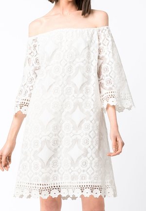 Femme portant une robe blanche en dentelle épaules dénudées avec des motifs floraux et géométriques et des manches cloche trois-quarts.