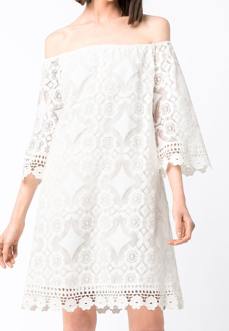Femme portant une robe blanche en dentelle épaules dénudées avec des motifs floraux et géométriques et des manches cloche trois-quarts.