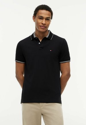 Ung mand iført en sort kortærmet polo med hvide kanter og beige bukser, stående med hænderne på ryggen mod en ensfarvet baggrund.
