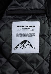 Melna iešūnu auduma, kuram ir balts uzlīme ar uzrakstu "PEGADOR OUTERWEAR." Uzlīme ietver kalnu grafiku un produkta apraksta tekstu.