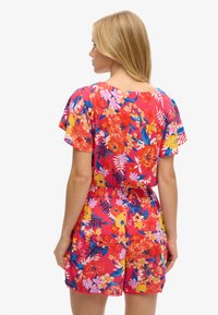 Salopette corta floreale con uno sfondo rosso vivace decorato da fiori rosa, gialli e blu. Tessuto leggero e arioso con una vestibilità rilassata.