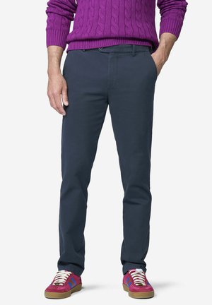 Mann trägt dunkelblaue Hose, einen lila Zopfmuster-Pullover und rote Sneakers mit blauen Streifen und weißen Schnürsenkeln, steht mit einer Hand in der Tasche.