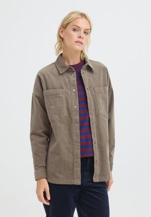 Blonde woman wearing an unbuttoned brown corduroy jacket over a blue and red striped shirt and dark pants, against a plain background.