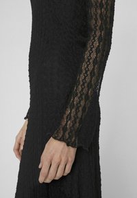 Melna mežģīņu kleita ar teksturētu augšdaļu un caurspīdīgām garām piedurknēm ar viļņotu apakšmalu, izceļot sarežģītus rakstus un slāņotu dizainu.