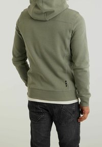 Felpa verde oliva con una finitura testurizzata, dotata di cappuccio, polsini a coste e un dettaglio di design a contrasto sul retro. Indossata sopra una camicia bianca.