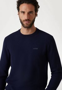 Maglione a maglia blu navy realizzato in materiale testurizzato, con scollo a girocollo e un sottile logo "GUESS" sul petto.