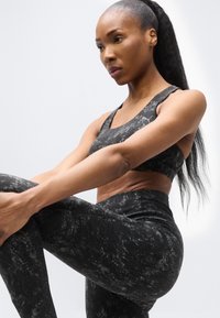 Γυμνασμένη γυναίκα με μαύρα αθλητικά ρούχα με σχέδια να τεντώνει το πόδι της, εστιάζοντας έντονα, με λευκό απλό φόντο.