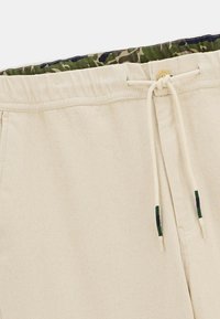Pantalons beiges avec une taille élastique à cordon de serrage, dotés d'un bouton et d'une doublure intérieure en camouflage vert et marron sur la taille.