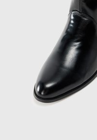 Cizma din piele neagră până la gleznă, cu vârful ascuțit, textură netedă, design minimalist și talpă subțire din cauciuc. Fără hardware vizibil sau modele.