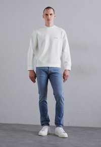 Valge see-sweater ribatud kaelusega, millel on väike logo. Ühendatud helesiniste teksade ja valgete tossudega hallil põrandal.
