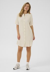 Robe beige à manches courtes, en tissu texturé, avec un ourlet arrondi. Porte avec des baskets grises et des chaussettes blanches à la cheville.