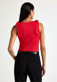 Červený top bez rukávů se mašlemi na ramenou, kombinovaný s černými kalhotami s texturovaným povrchem a úzkým černým páskem.