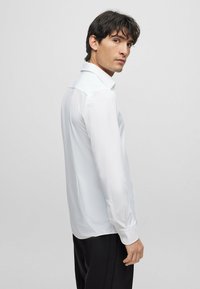 Camisa blanca de manga larga con un diseño liso, textura suave y un cuello clásico. Presenta puños abotonados y un ajuste entallado.