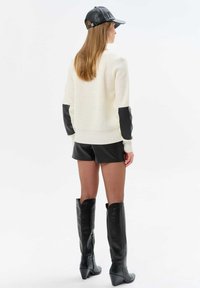 White knit sweater with black leather sleeves, paired with black leather shorts and knee-high boots, featuring a tapered heel and pointed toe.