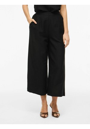 Žena nosiace čierne široké zúžené skrátené nohavice s vysokým pásom a hnedé lodičky so špicatou špičkou, stojaca s jednou rukou v vrecku.