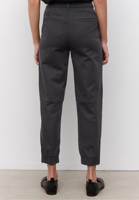 Henkilö, jolla on päällään mustat lyhyet housut, joissa on joustavat hihnat, ja mustat liukuhalkaisijat, seisoo yksinkertaista valkoista seinää vasten.