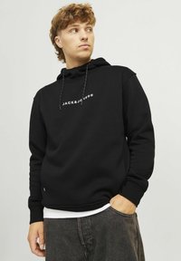 Crna dukserica od mekog pamuka, s bijelim logotipom "JACK & JONES" preko prsa, s kapuljačom i opuštenim krojem.