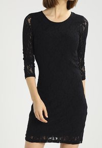 Robe en dentelle noire à manches trois-quarts, présentant un motif floral et une coupe ajustée. Le tissu a une texture douce et un léger éclat.