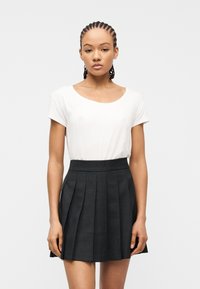 Νεαρή γυναίκα με πλεγμένα μαλλιά, φορώντας ένα λευκό κοντομάνικο τοπ και μια μαύρη πλισέ φούστα με ψηλή μέση, στέκεται μπροστά σε ένα απλό ανοιχτό φόντο.