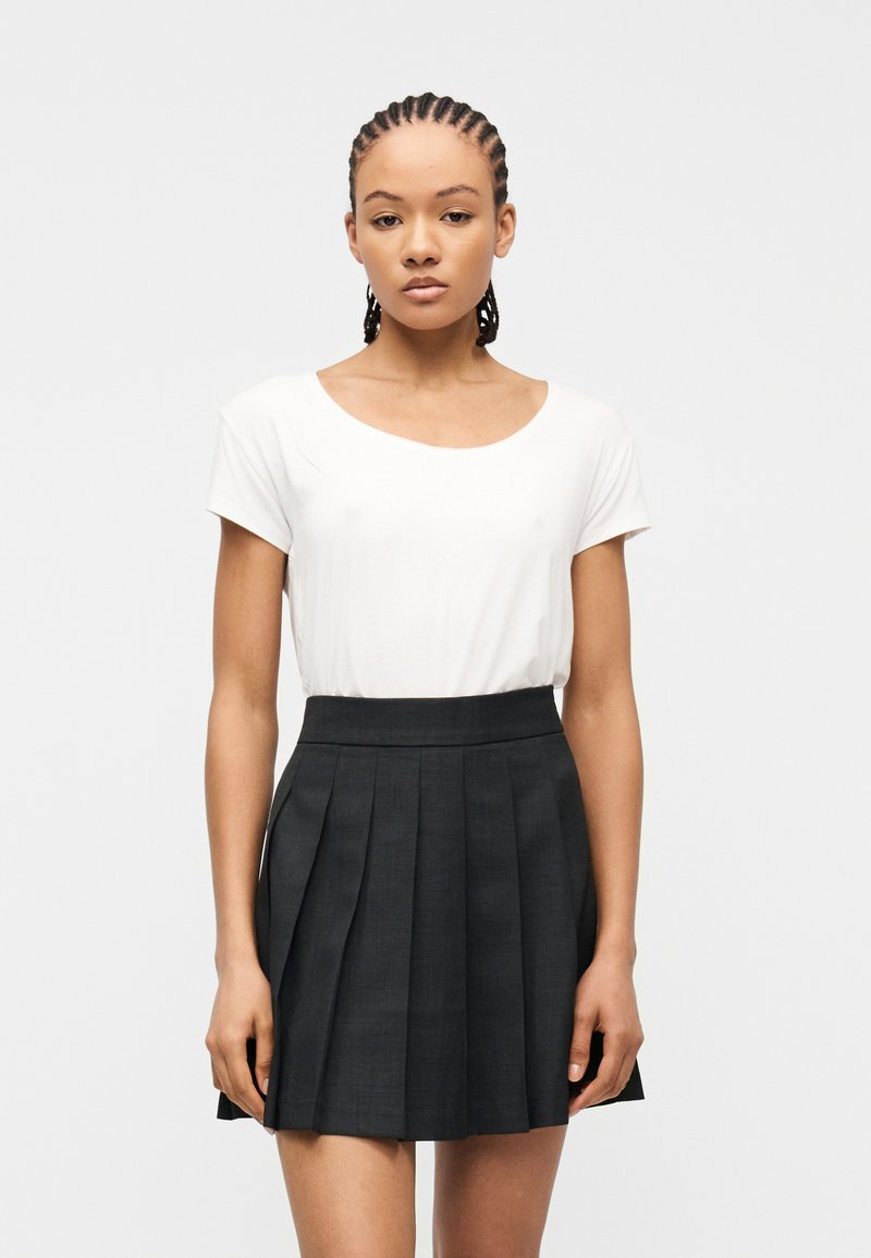 Νεαρή γυναίκα με πλεγμένα μαλλιά, φορώντας ένα λευκό κοντομάνικο τοπ και μια μαύρη πλισέ φούστα με ψηλή μέση, στέκεται μπροστά σε ένα απλό ανοιχτό φόντο.