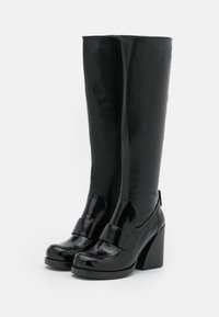 Bottes noires hautes en cuir synthétique brillant, avec un talon bloc, un bout arrondi et un détail nœud enroulé autour de la cheville.