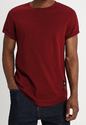 Homme portant un t-shirt uni à manches courtes rouge foncé avec les manches retroussées et un jean bleu foncé, debout avec une main dans la poche.