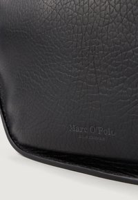 Sac en cuir noir avec une surface texturée. Présente un logo en relief "Marc O'Polo" et une bordure lisse. Design minimaliste, forme rectangulaire.