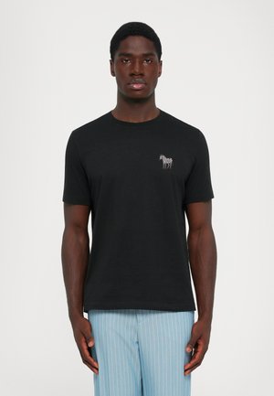 Ung man som bär en svart t-shirt med en liten zebra-logotyp på bröstet och ljusblå randiga byxor, stående mot en vit bakgrund.