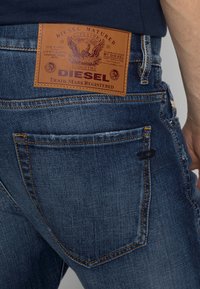 Denim jeans in dark blue with a textured finish, featuring a brown leather patch on the back with embossed logo and eagle design.