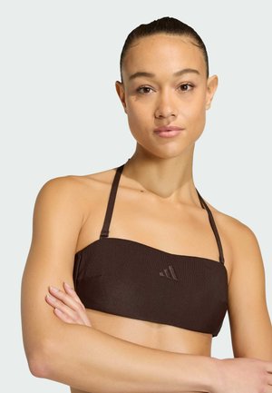 Młoda kobieta z założonymi rękami, ubrana w ciemnobrązowy prążkowany top bikini typu halter na jednolitym jasnym tle.