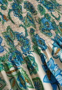 Gros plan sur un tissu crème avec un imprimé floral bleu, vert et jaune et une couture smock élastique créant une texture froncée.