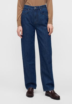 Person trägt dunkelblaue Jeans mit geradem Bein, ein braunes Langarmshirt und dunkelbraune Loafers und steht vor einem einfarbigen Hintergrund.