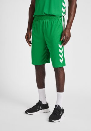 Grønne sportsshorts med elastisk talje, med hvide chevron-detaljer på siderne, parret med sorte sneakers og hvide sokker.