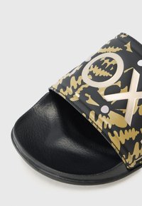 Svart slidesandaler med en blank overflate som har et gullfarget abstrakt mønster og hvite detaljer, flat såle og et bredt åpent tådesign.