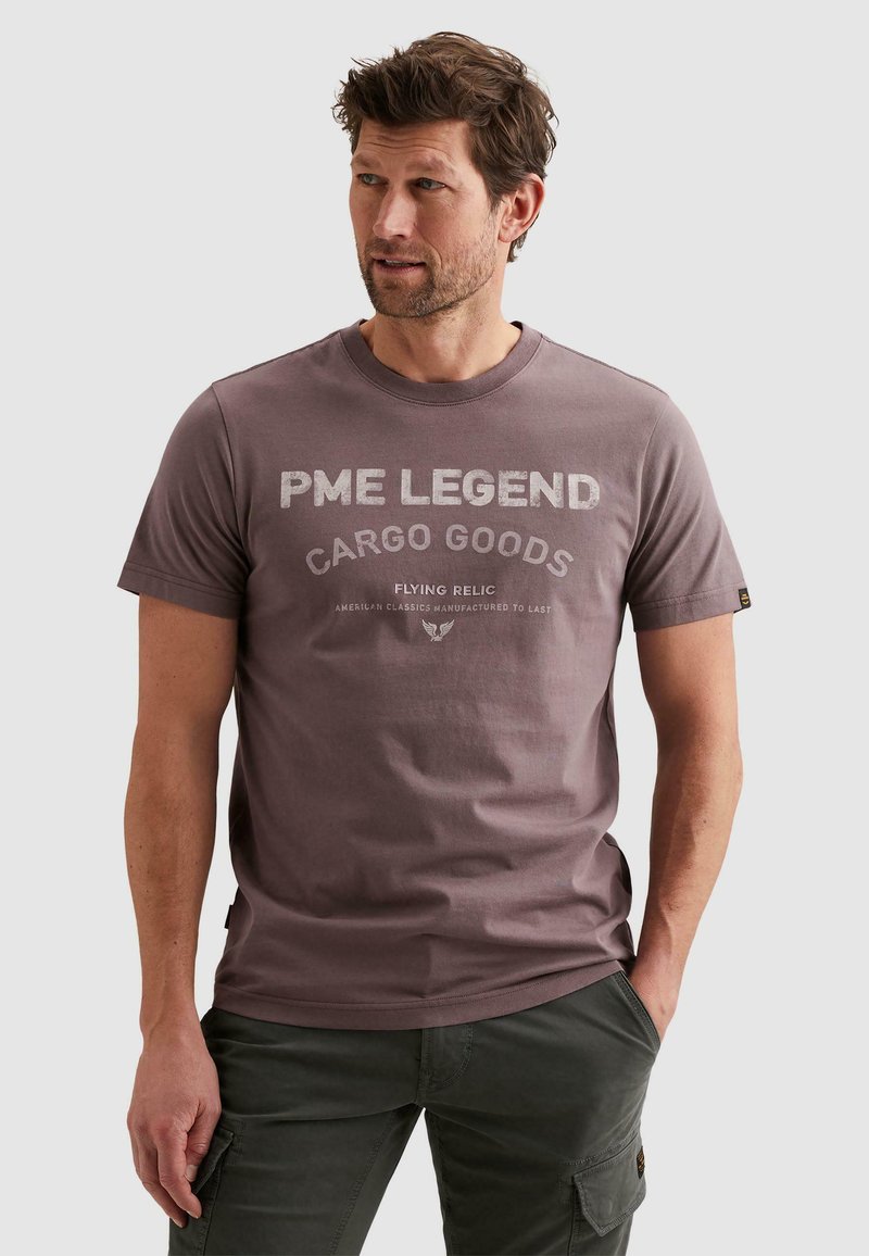 Homme aux cheveux bruns courts portant un t-shirt graphique mauve PME Legend et un pantalon cargo foncé, debout avec une main dans la poche.