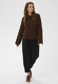 Brown cable-knit sweater with a textured pattern, wide sleeves, paired with loose black trousers and brown shoes. Neutral background.