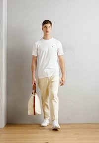Hvid kortærmet t-shirt, cremefarvede bukser og hvide sneakers, bærende en cremefarvet taske med brune håndtag, stående på et trægulv mod en grå væg.