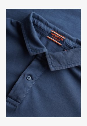 Ciemnoniebieska koszulka polo z teksturowanej tkaniny. Wyposażona w klasyczny kołnierzyk, zapięcie na dwa guziki i metkę marki w kolorze czerwonym i pomarańczowym.