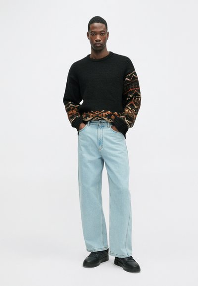 Fekete kötött pulóver mintás narancssárga és barna ujjakkal, világoskék, laza szabású farmernadrággal és fekete cipővel párosítva. Egyszínű fehér háttér.