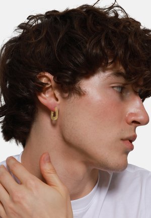Gold hoop earring displayed. The earring has a geometric, open design with a smooth metallic surface. The model has curly dark hair and wears a white shirt.