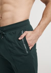 Donkergroene joggerbroeken van zacht materiaal, met een tailleband met trekkoord en een zijzak met witte "Jockey" branding.