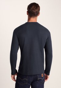 Man wearing a long-sleeve dark textured shirt and dark jeans, shown from the back against a plain light background.